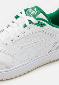 Basket en cuir synthétique blanc avec des accents verts, dotée de lacets plats blancs et d'une semelle en caoutchouc texturée. Logo Puma embossé sur le côté.