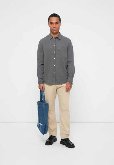 Chemise gris à boutons avec une poche poitrine, pantalon couleur crème et sac fourre-tout en tissu bleu. Le modèle se tient devant un fond blanc uni.
