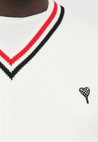 Baltas megztinis su giliais V formos iškirpte, puoštas raudonais ir juodais dryžiais palei iškirptę bei mažyliu juodu širdies logotipu ant krūtinės.