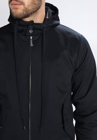 Black zip-up jacket with a hood, featuring a plaid lining, drawstrings, and button details on the pockets. Smooth, textured material.