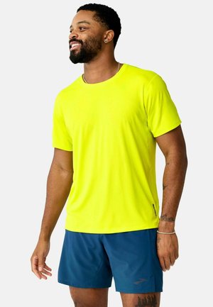 Uomo sorridente, indossa una camicia gialla brillante a maniche corte e pantaloncini sportivi blu navy, in piedi davanti a uno sfondo semplice.