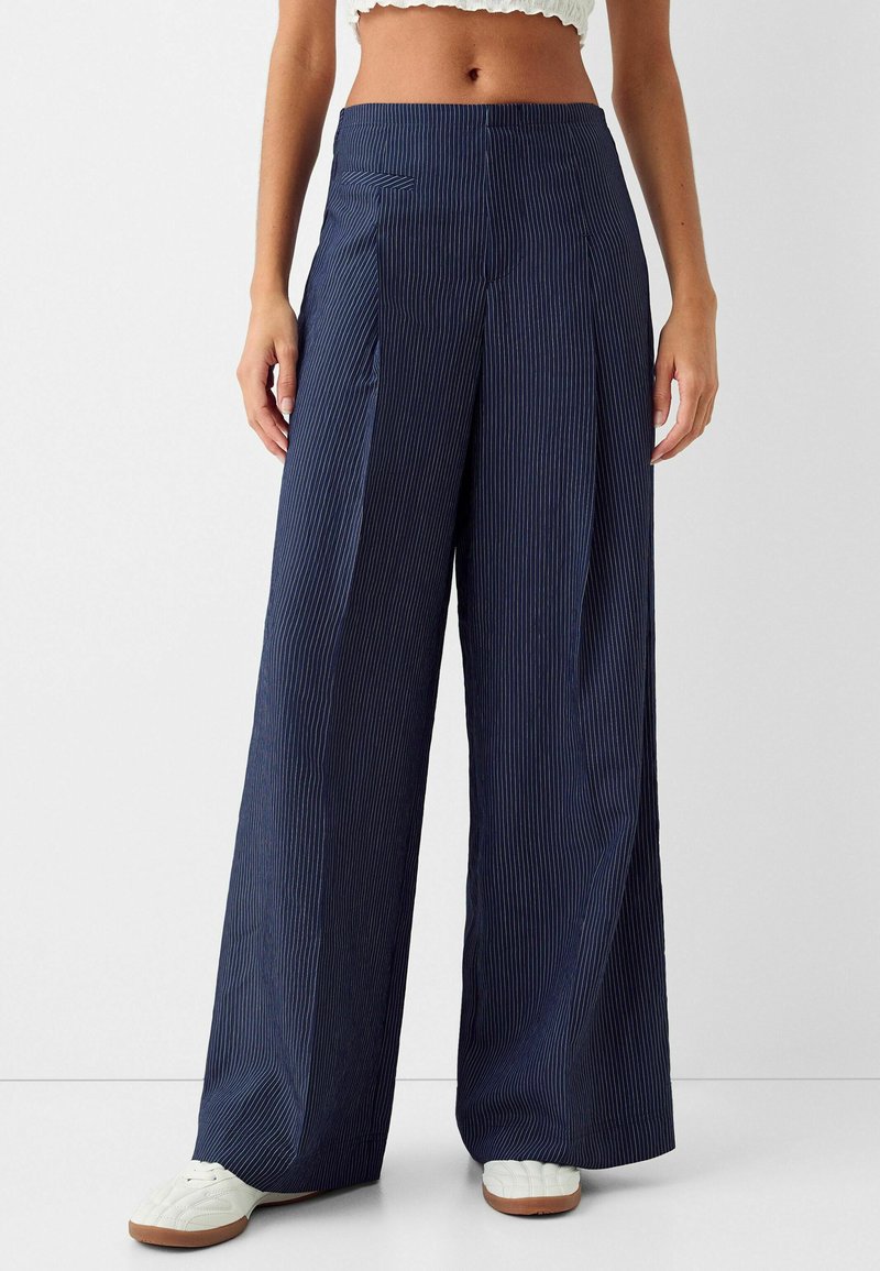 Pantaloni a gamba larga di colore blu navy realizzati in tessuto leggero con righe verticali bianche. Presentano una vita alta e pieghe frontali, abbinati a sneakers bianche.