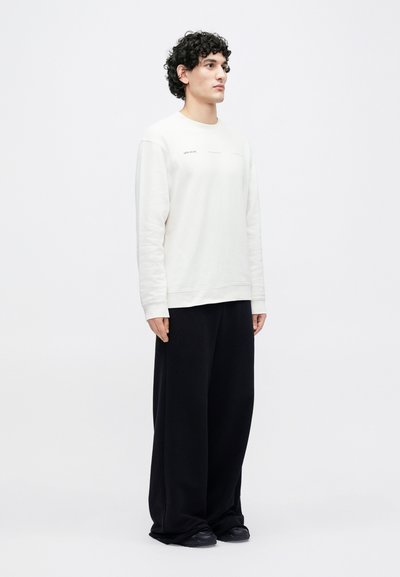 Krémová mikina s okrúhlym výstrihom, dlhými rukávmi a minimálnym textom na prednej strane, doplnená voľnými čiernymi nohavicami a tmavými topánkami.