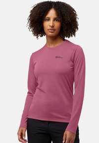 Frau mit lockigem Haar, die ein langärmliges mauvefarbenes Jack Wolfskin-Hemd und schwarze Hosen trägt, steht vor einem hellen Hintergrund.