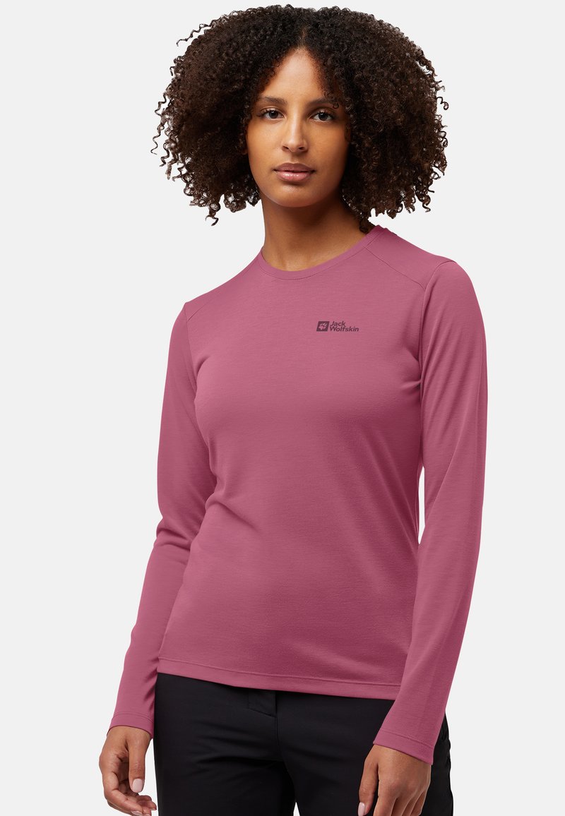 Frau mit lockigem Haar, die ein langärmliges mauvefarbenes Jack Wolfskin-Hemd und schwarze Hosen trägt, steht vor einem hellen Hintergrund.