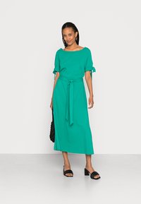 Vestido midi verde-água com mangas curtas e detalhes de laço, apresentando um cinto na cintura. Modelo usa sandálias pretas de salto aberto e carrega uma bolsa preta.