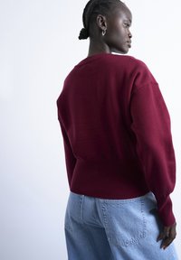 Femme aux cheveux tressés portant un pull rouge foncé et un jean en denim bleu clair, debout de profil devant un fond clair uni.