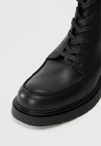 Bota de cuero negro texturizado con cordones, suela gruesa y diseño de punta mocasin frente a un fondo claro y liso.
