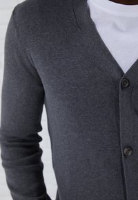 Primer plano de una persona que lleva un cárdigan gris oscuro abotonado sobre una camiseta blanca de cuello redondo, mostrando el torso y el brazo izquierdo.