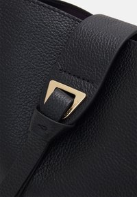 Dettaglio di una borsa in pelle nera con finitura testurizzata, una tracolla spessa e un accent hardware triangolare in tono oro.