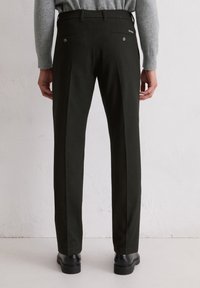 Pantalons noirs ajustés avec une texture lisse, devant plissé et deux poches arrière. Portés avec un pull gris et des chaussures noires.