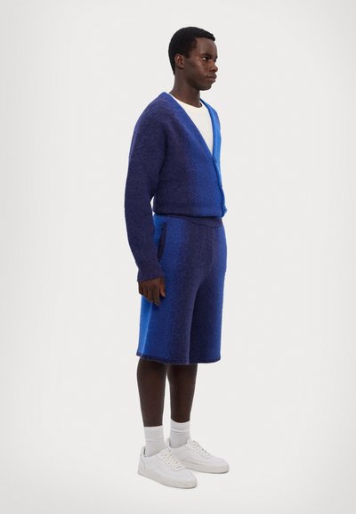 Strikket blå cardigan og matchende shorts med gradient-design. Hvid t-shirt under, parret med hvide sneakers og sokker.