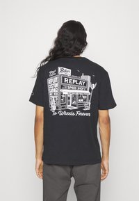 Černé bavlněné tričko s bílým grafickým potiskem zobrazujícím motocyklovou dílnu a textem "Na kolech navždy" na zadní straně.