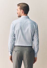 Uomo visto di spalle che indossa una camicia a maniche lunghe a quadri azzurri e pantaloni eleganti grigi, su uno sfondo chiaro e uniforme.