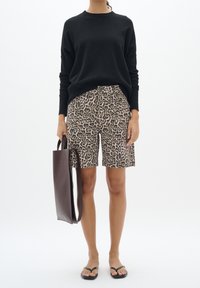 Femme portant un haut noir à manches longues, un short léopard jusqu'aux genoux, des tongs noires, et tenant un sac cabas en cuir marron foncé.