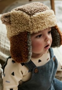 Textured hat in beige, brown, and gray with ear flaps. Worn by a child, complemented by a patterned shirt and blue overalls.