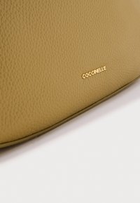 Sac en cuir beige texturé avec des bords arrondis et un logo doré "Coccinelle" bien visible.