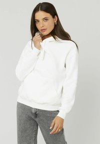 Sweatshirt à capuche blanche avec cordon de serrage, poche kangourou et poignets côtelés, associé à un jean gris. Texture douce, design décontracté.