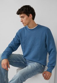 Niebieski sweter o fakturowanej strukturze z plecionym wzorem, okrągłym dekoltem i prążkowanymi mankietami; w połączeniu z jasnoniebieskimi jeansami, siedzący na neutralnym tle.