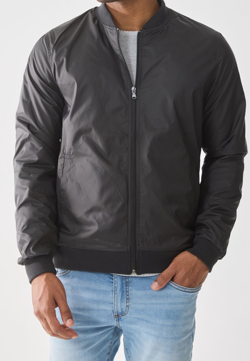 Homme portant une veste bomber noire zippée sur une chemise grise et un jean bleu clair, une main dans une poche et l'autre détendue.