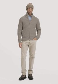 Beige gestrickte Zip-Up-Jacke mit einem strukturierten Muster, kombiniert mit einem hellblauen Hemd, beigen Hosen und braunen Schnürstiefeln.