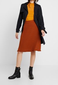 Svart trenchcoat med knapp detaljer, orange ribbad tröja, texturerad rostfärgad kjol med volangkant, och svarta ankelstövlar med sidogetningar.