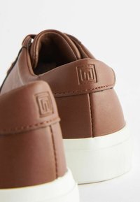 Brune syntetiske lædersneakers med hvide gummisåler. Har logo prægede på hælen og et strømlinet design. Teksturdetaljer på overfladen.