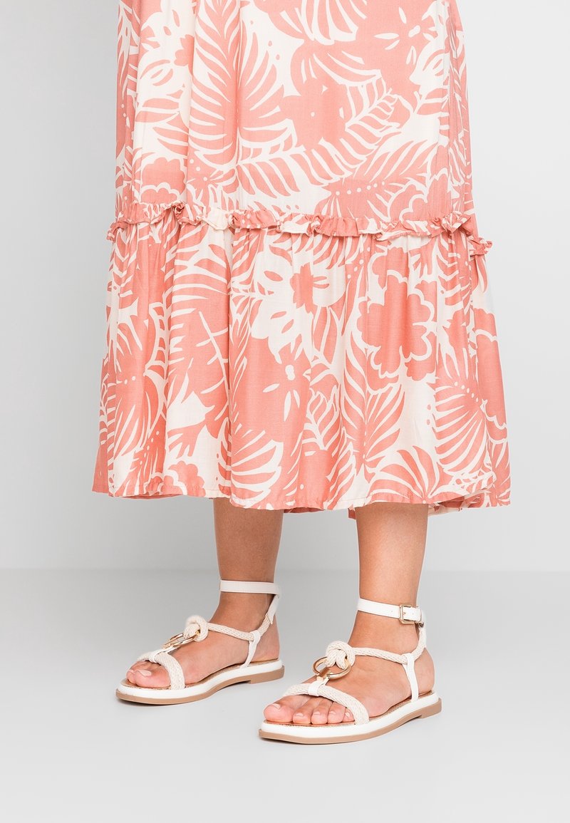 Bas des jambes portant des sandales blanches à brides tressées avec des détails d'anneaux dorés, associées à une jupe midi fleurie pêche et blanche.