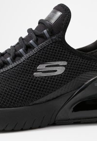 Czarne buty sportowe z siateczkowej cholewki, teksturowanymi akcentami, gumową podeszwą i metalicznym logo. Sznurowany design z wzmocnionym noskiem.