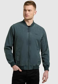 Jeune homme portant une veste bomber vert foncé avec fermeture éclair à l'avant et poignets côtelés, debout avec une main dans la poche, regardant vers sa gauche.