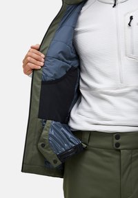 Groene buitenjas met een gewatteerde blauwe binnenkant, zwarte zak en drukknoopsluitingen. Gedragen over een witte gestructureerde top en groene broek.