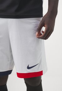 Baltie sporta šorti ar vertikālo tekstūru, tumši zilo akcentu un sarkanu apakšējo malu. Nike logo tumši zilā krāsā kreisajā pusē.