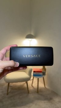 Main tenant une étui à lunettes Versace rectangulaire noir avec un fauteuil beige, une table d'appoint, des livres et une lampe en arrière-plan.