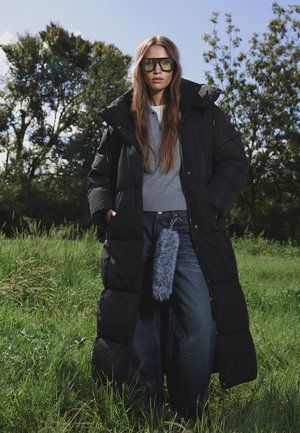 Lange zwarte pufferjas met een gestructureerde afwerking, grijze gebreide trui en wijde spijkerbroek. Accessoires omvatten een fluffy grijze sleutelhanger.