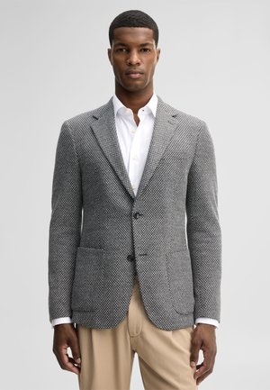 Man die een grijs patroonblazer draagt over een wit overhemd met knopen en beige broek, staand met het gezicht naar voren tegen een effen achtergrond.