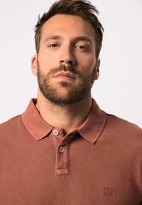 Polo en coton brun avec un col à boutons, arborant un logo sur la poitrine gauche et un tissu texturé, exposé sur un fond neutre.