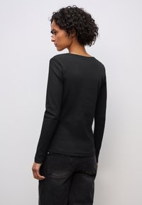 Schwarzes Langarmoberteil aus geripptem Material mit runder Ausschnitt. Der Rücken ist figurbetont und wird mit dunklen Jeans kombiniert. Minimalistisches Design.