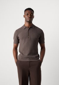 Polo de punto marrón con mangas cortas, textura acanalada y tapeta de tres botones. Diseño ajustado combinado con pantalones marrones oscuros.