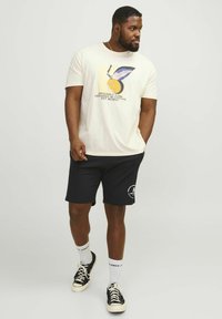 Una maglietta grafica color crema presenta una stampa vivace con un design a cerchio e foglia. Indossata con pantaloni corti neri e sneakers nere con dettagli bianchi.