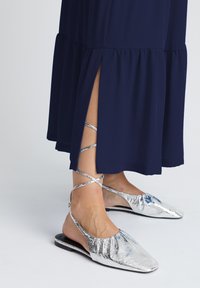 Silberne metallic flache Schuhe mit gerafftem Design, Knöchelriemen und spitzer Zehenpartie; getragen zu einem marineblauen, plissierten Kleid mit seitlichen Schlitzen.