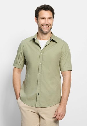 Lächelnder Mann mit kurzen lockigen Haaren und Bart, trägt ein hellgrünes kurzärmeliges Hemd und beige Hose, Hände teilweise in den Taschen.