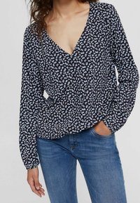 Donna che indossa una blusa nera a maniche lunghe con piccoli motivi floreali bianchi e jeans blu, con una mano in tasca, che sta in piedi contro uno sfondo semplice.