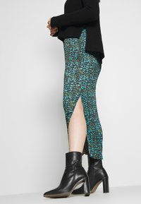 Svart långärmad tröja i kombination med en midjefärgad kjol med ett blått och grönt abstrakt mönster, sidöppning och svarta ankelskor.