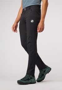 Zwarte outdoor broeken van flexibele stof, met zijzakken met rits en een logo patch. Gemaakt in combinatie met groene wandelschoenen en een blauwe shirt.
