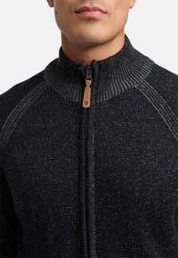 Sort strikket sweater med høj krave, der har en kontrasterende lynlås og en lædertrækstrop. Struktureret mønster synligt overalt.