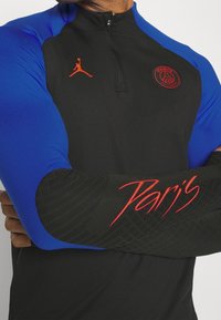 Schwarzes Quarter-Zip-Oberteil mit blauen Ärmeln, das ein rotes Jordan-Logo, ein Paris-Wappen und "Paris" in roter Schreibschrift auf dem strukturierten Ärmel zeigt.