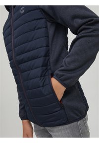 Veste bleu marine avec un rembourrage matelassé sur le devant, des manches en tissu lisse, des poches latérales et une fermeture éclair. Coutures contrastées visibles.