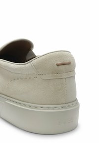 Ljusbeige mocka slip-on sko med en strukturerad yta, utrustad med gummisula och en minimalistisk brun detalj på hälen.