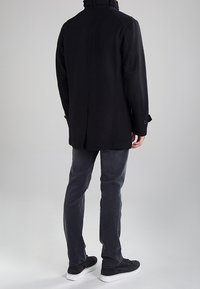 Cappotto di lana nero con colletto alto, dettagli con bottoni e vestibilità su misura, abbinato a pantaloni grigio scuro e sneakers nere.
