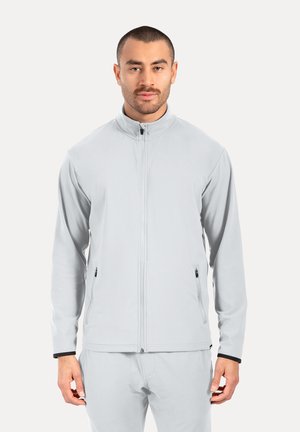Mann trägt eine hellgraue Jacke mit Reißverschluss und passende Hosen, steht mit Blick nach vorne vor einem schlichten hellen Hintergrund.